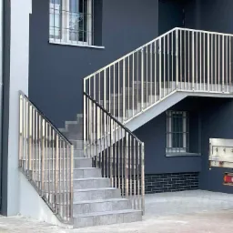 Zewnętrzne schody z szarymi płytkami i nowoczesną balustradą ze stali nierdzewnej, prowadzące do ciemnoszarego budynku z oknami z kratami, widok z boku z fragmentem przyczepy samochodowej w tle.