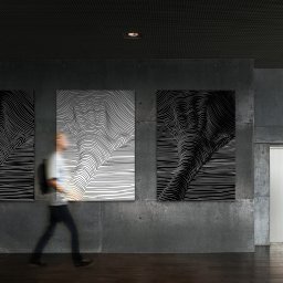Julia Bobrukiewicz - Trzy abstrakcyjne obrazy z czarno-białymi liniami na betonowej ścianie w korytarzu, obok windy, z rozmytą sylwetką przechodzącej osoby.