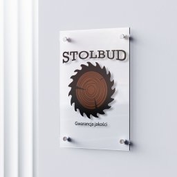 Julia Bobrukiewicz - Logo firmy Stolbud na przezroczystej tablicy przymocowanej do ściany. Logo przedstawia piłę tarczową z przekrojem drewna w środku i napis 'Gwarancja jakości' pod spodem.