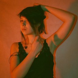 Julia Bobrukiewicz - Portret kobiety w czarnej bieliźnie z koronką, oświetlonej pomarańczowym i zielonym światłem, z ręką uniesioną do włosów i tatuażem serca na dekolcie.