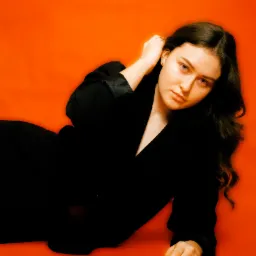 Portret kobiety w czarnej marynarce na pomarańczowym tle, z ręką uniesioną do włosów.