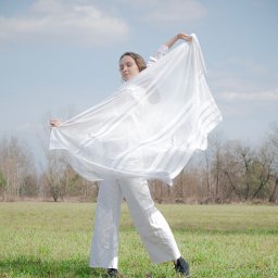 Julia Bobrukiewicz - Kobieta w białym stroju, trzymająca w dłoniach zwiewną, białą tkaninę na tle zielonej łąki i błękitnego nieba z chmurami.