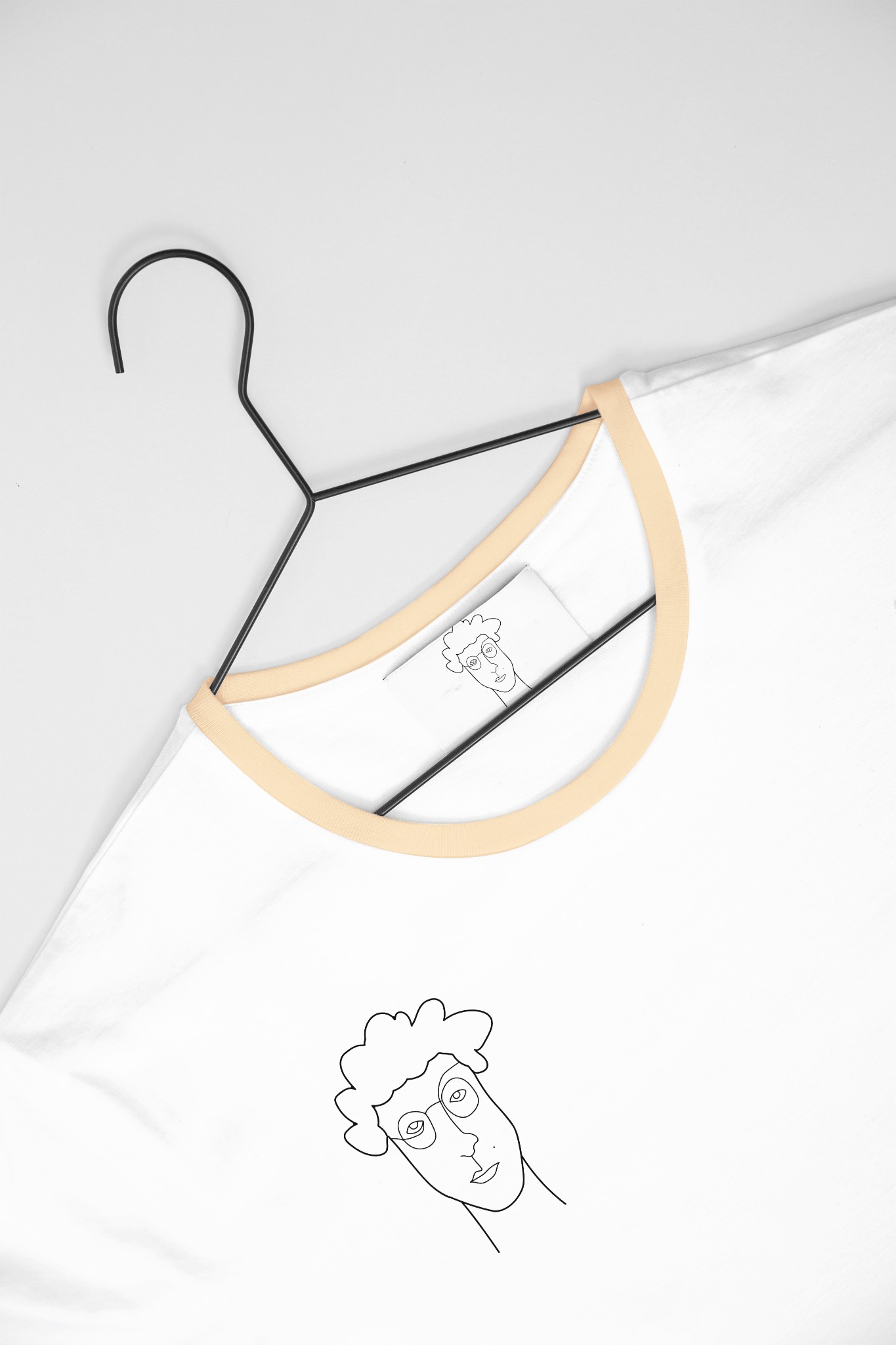 Biała koszulka z czarnym konturowym rysunkiem twarzy na wieszaku, wykonana w Kobyłce. Rysunek powtórzony na metce.