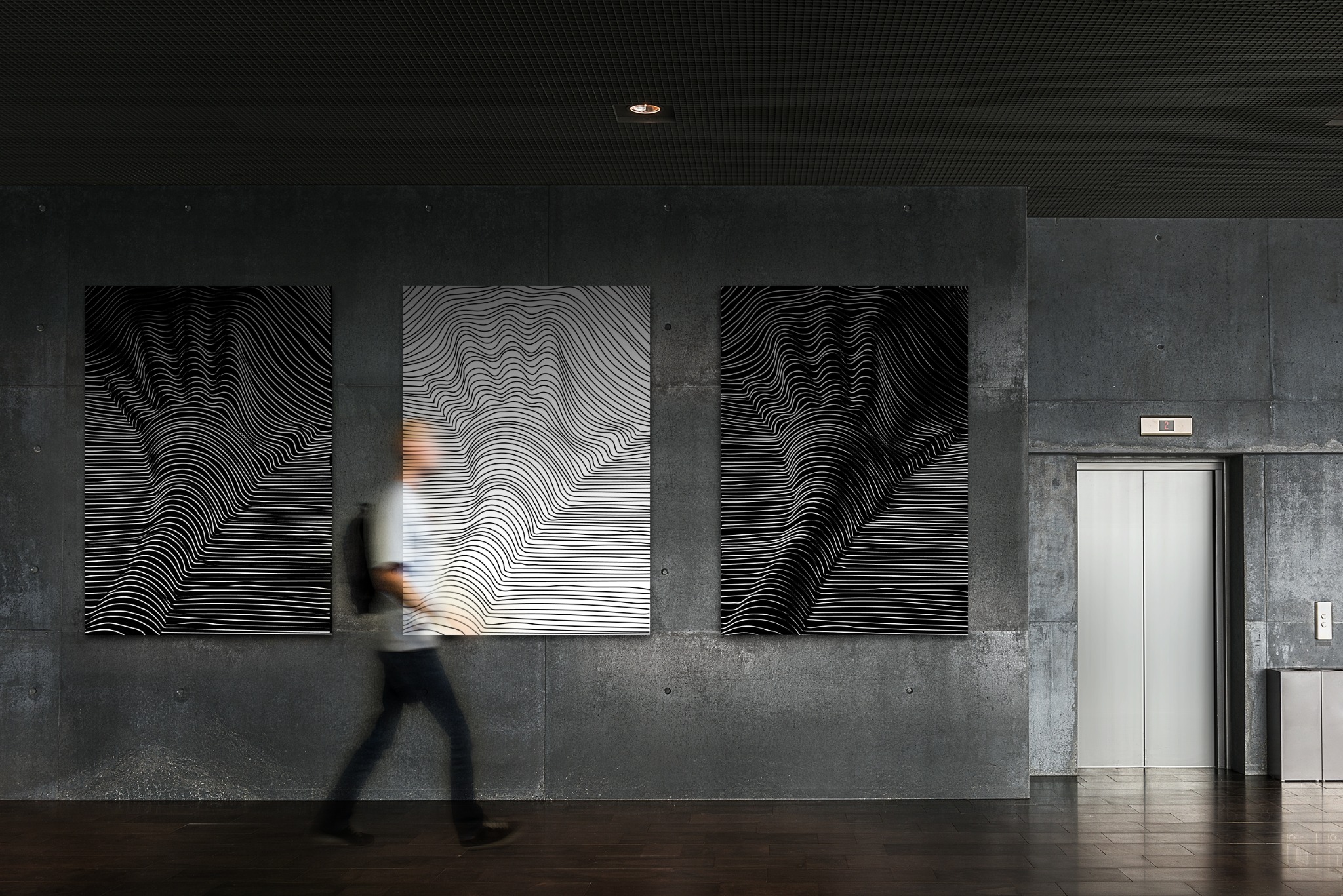 Trzy abstrakcyjne obrazy z czarno-białymi liniami na betonowej ścianie w korytarzu, obok windy, z rozmytą sylwetką przechodzącej osoby.
