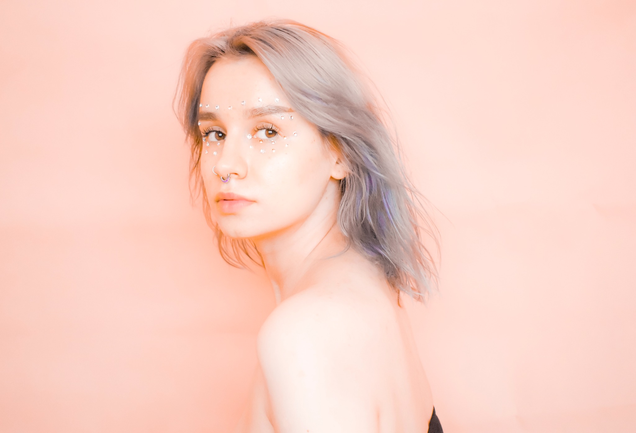 Portret kobiety z szarymi włosami i kryształkami na twarzy, patrzącej przez ramię na jasnoróżowym tle.