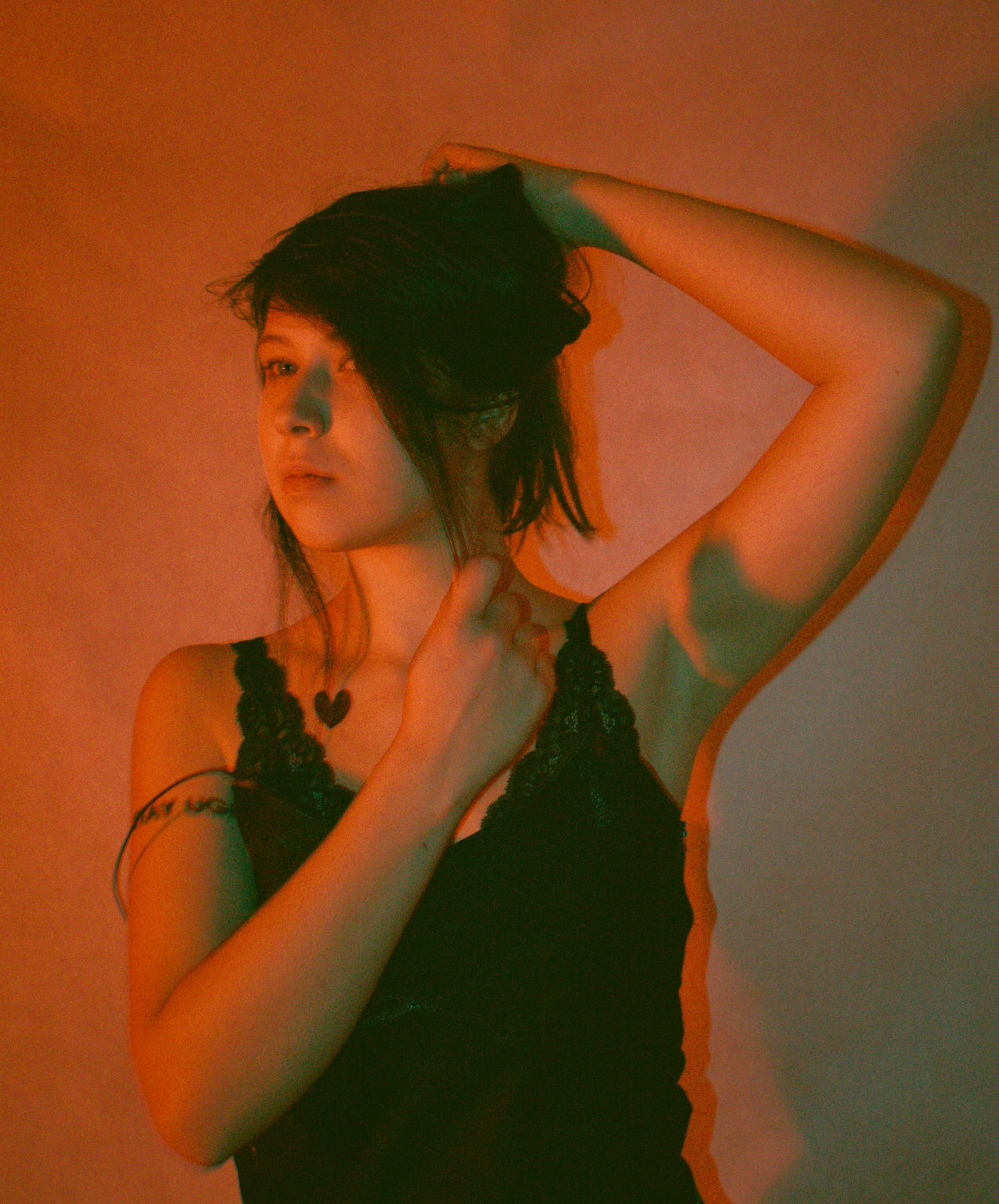 Portret kobiety w czarnej bieliźnie z koronką, oświetlonej pomarańczowym i zielonym światłem, z ręką uniesioną do włosów i tatuażem serca na dekolcie.