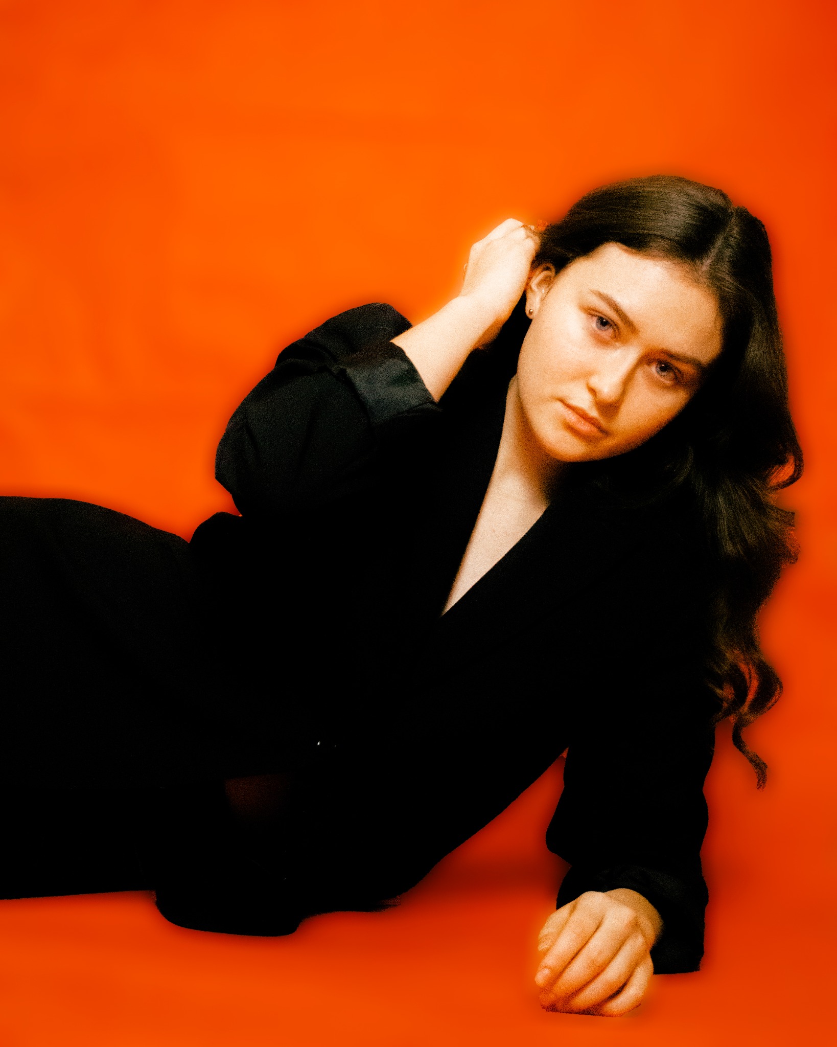 Portret kobiety w czarnej marynarce na pomarańczowym tle, z ręką uniesioną do włosów.