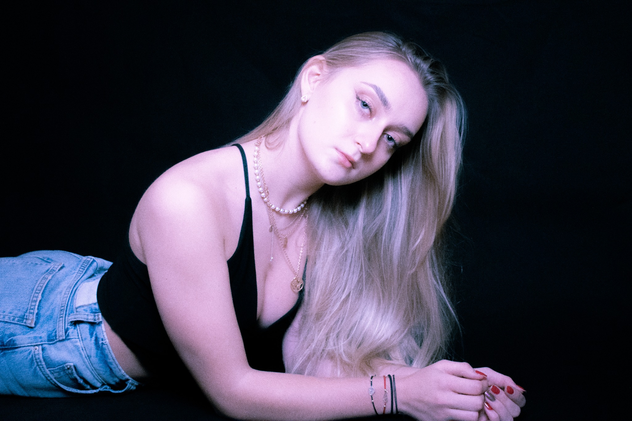 Portret leżącej blondynki w dżinsach i czarnym topie, z kilkoma naszyjnikami i bransoletkami na tle ciemnego tła.