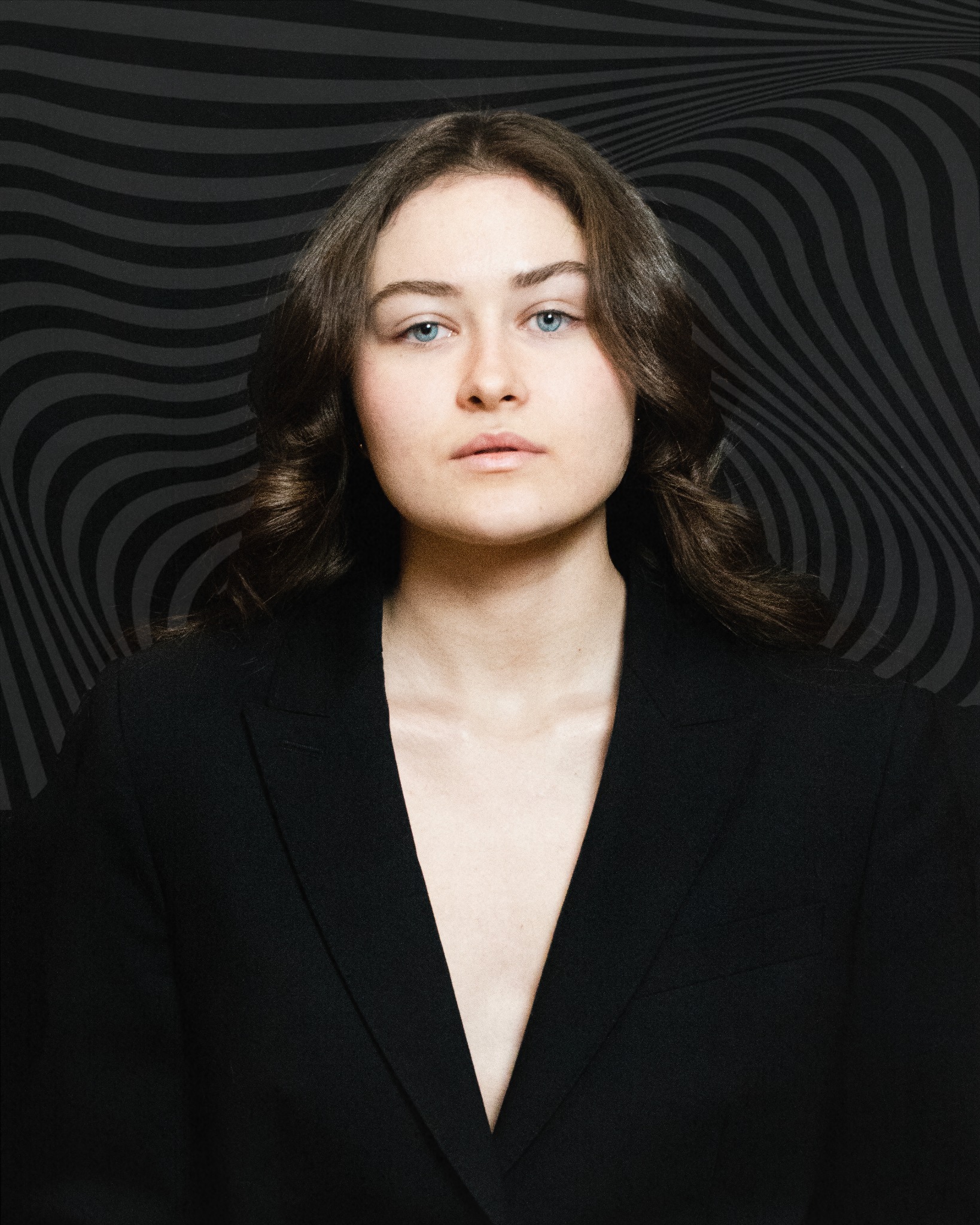 Portret kobiety z niebieskimi oczami w czarnej marynarce na tle abstrakcyjnego, falistego wzoru w odcieniach szarości, patrzącej prosto w obiektyw.