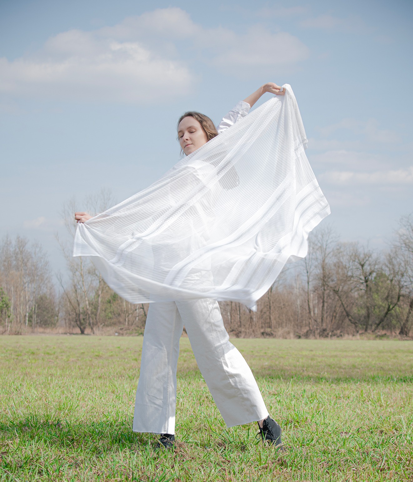 Kobieta w białym stroju, trzymająca w dłoniach zwiewną, białą tkaninę na tle zielonej łąki i błękitnego nieba z chmurami.