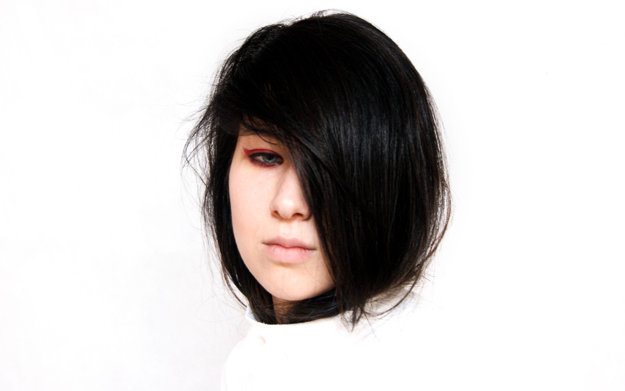 Portret kobiety z czarnymi włosami zasłaniającymi część twarzy i z delikatnym, czerwonym makijażem oczu na białym tle.