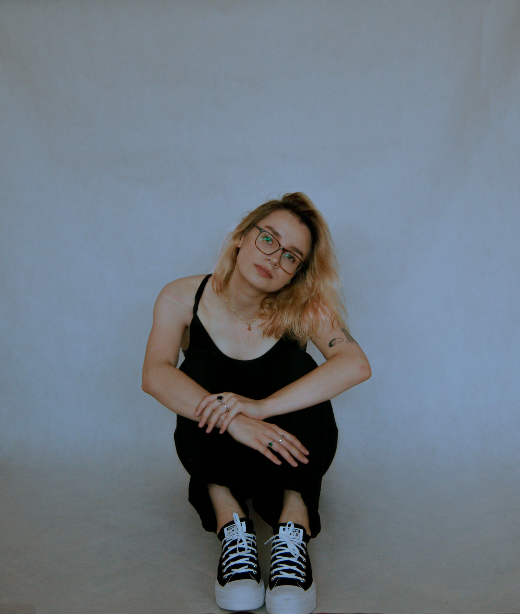 Portret młodej kobiety w okularach, siedzącej na kucki, z rękami splecionymi na kolanach, na jednolitym, jasnoszarym tle.
