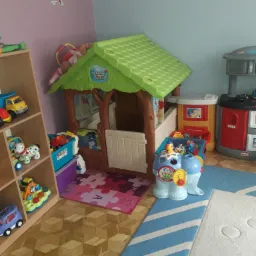 Pokój zabaw dla dzieci z drewnianą podłogą, regałem z zabawkami, domkiem z motywem Kubusia Puchatka, kuchnią zabawkową i dywanem w paski.