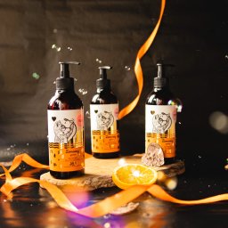 Martin Błaszkiewicz - Trzy butelki brązowego żelu pod prysznic z etykietami z rysunkiem lisa, umieszczone na drewnianej desce, otoczone pomarańczową wstążką, plasterkiem pomarańczy i bąbelkami na ciemnym tle.