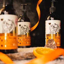 Martin Błaszkiewicz - Trzy butelki żelu pod prysznic z pompką, z etykietami przedstawiającymi rysunkowego lisa, ustawione na drewnianej desce posypanej białym proszkiem, z plasterkiem pomarańczy i pomarańczową wstążką.