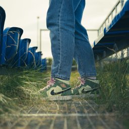 Martin Błaszkiewicz - Osoba w dżinsach i zielonych butach sportowych Air Jordan z różowymi sznurówkami stoi na metalowej kratce w otoczeniu trawy i rzędów niebieskich siedzeń, Wrocław.