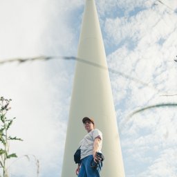 Martin Błaszkiewicz - Osoba z aparatem stoi u podstawy turbiny wiatrowej, spoglądając w dół, z trawą i chwastami na pierwszym planie.