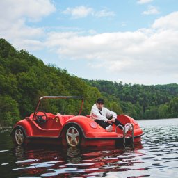 Martin Błaszkiewicz - Młody mężczyzna w białej bluzie i czarnej czapce siedzi w czerwonym, samochodopodobnym rowerze wodnym na jeziorze, zalesione wzgórze w tle i błękitne niebo z białymi obłokami.