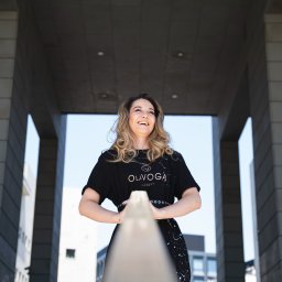 Martin Błaszkiewicz - Uśmiechnięta kobieta w czarnej sukience z logo marki, stojąca na schodach z metalową barierką, w tle nowoczesna architektura miejska i błękitne niebo.