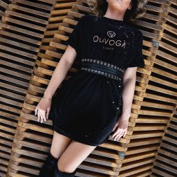 Martin Błaszkiewicz - Kobieta w czarnej sukience z logo leży na drewnianej konstrukcji, pozując do zdjęcia z uniesioną głową.