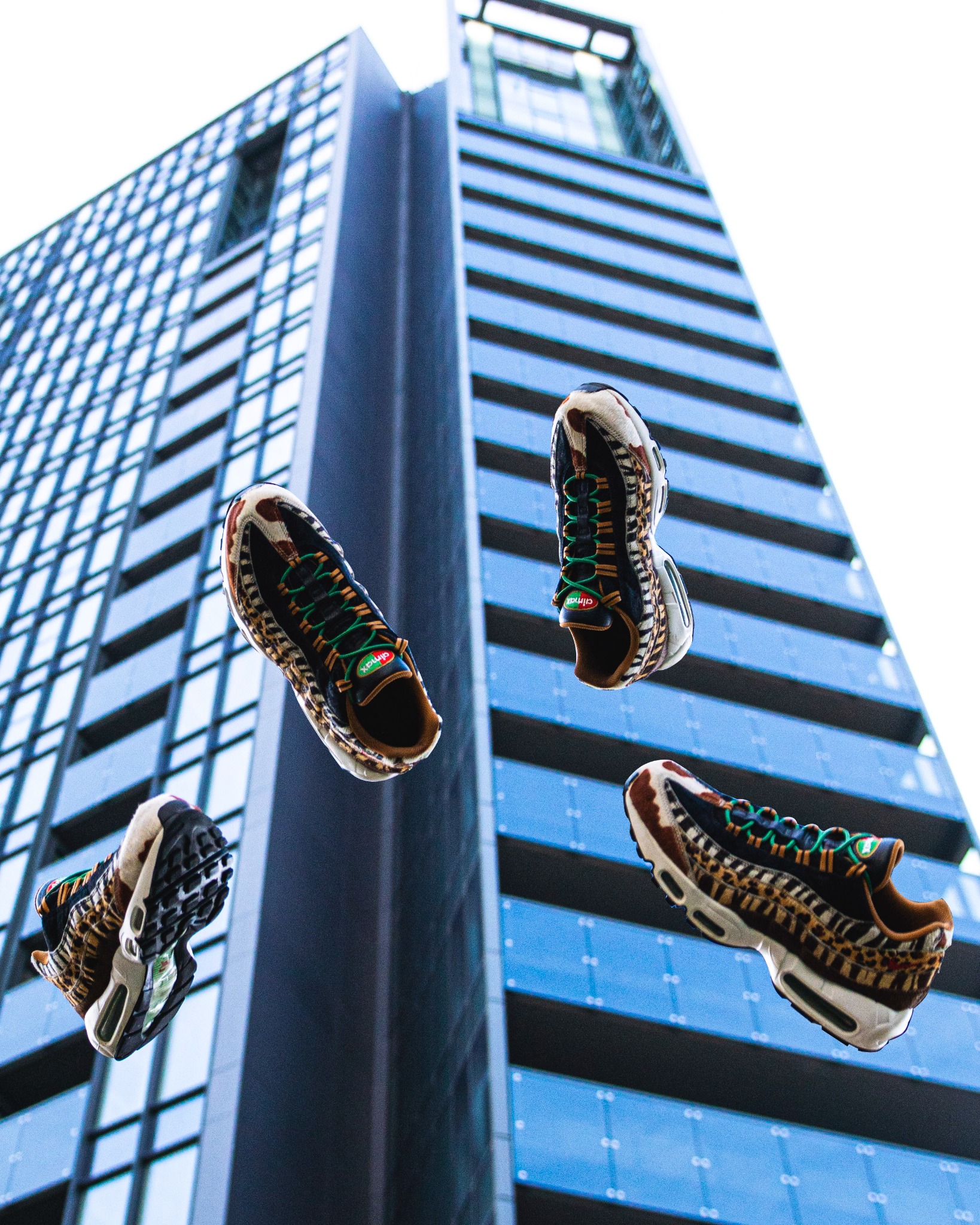 Trzy buty sportowe z motywem zwierzęcym unoszą się w powietrzu na tle nowoczesnego, wysokiego budynku z niebieskimi oknami.