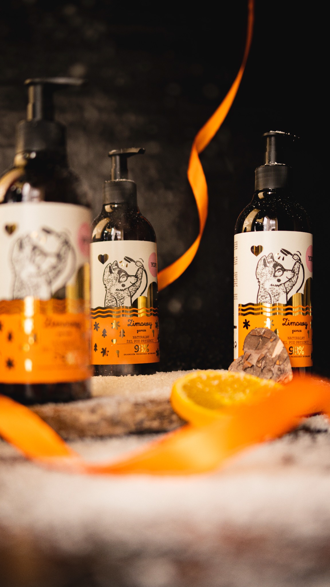 Trzy butelki żelu pod prysznic z pompką, z etykietami przedstawiającymi rysunkowego lisa, ustawione na drewnianej desce posypanej białym proszkiem, z plasterkiem pomarańczy i pomarańczową wstążką.