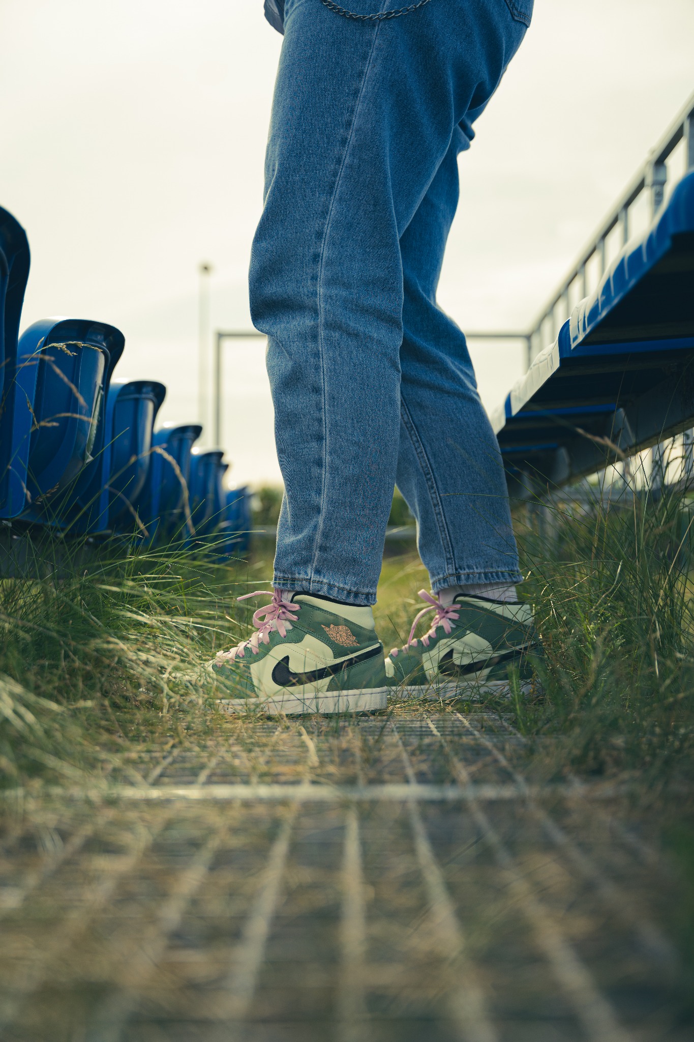 Osoba w dżinsach i zielonych butach sportowych Air Jordan z różowymi sznurówkami stoi na metalowej kratce w otoczeniu trawy i rzędów niebieskich siedzeń, Wrocław.
