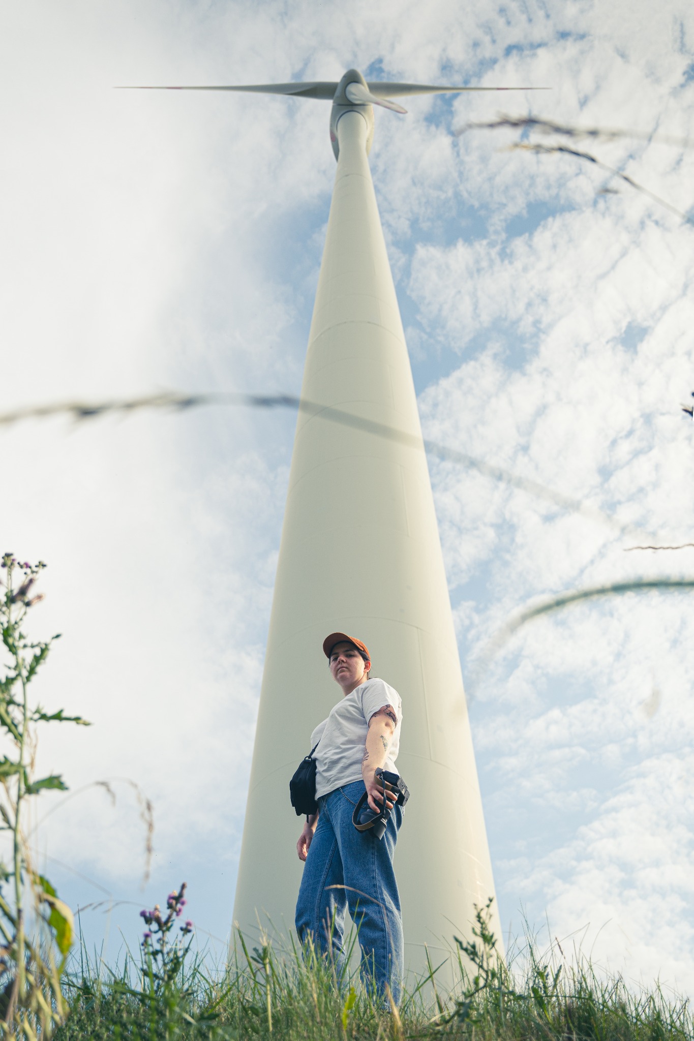 Osoba z aparatem stoi u podstawy turbiny wiatrowej, spoglądając w dół, z trawą i chwastami na pierwszym planie.