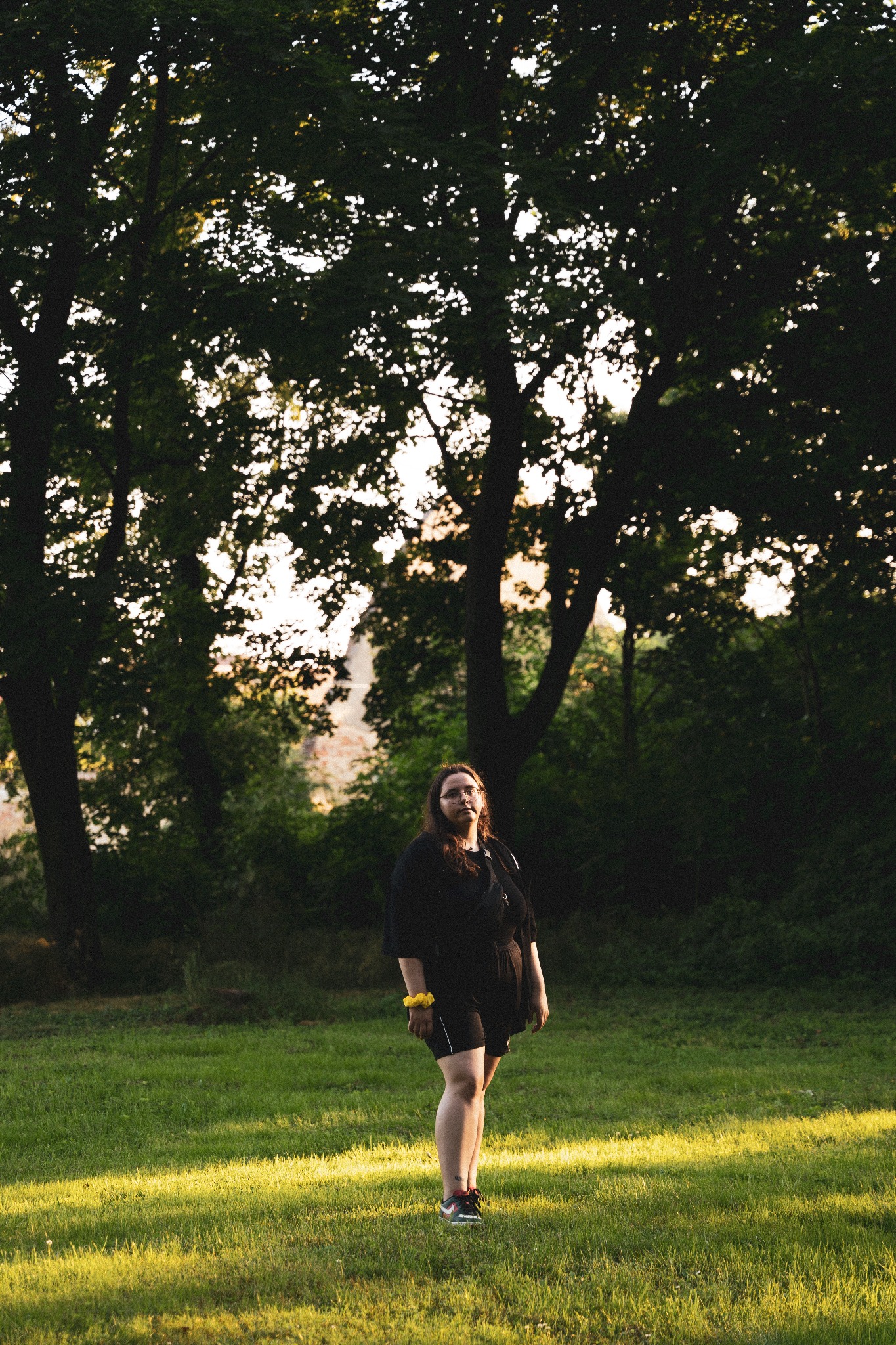 Portret kobiety w czarnym stroju na tle parku z drzewami, z promieniem słońca padającym na trawę.