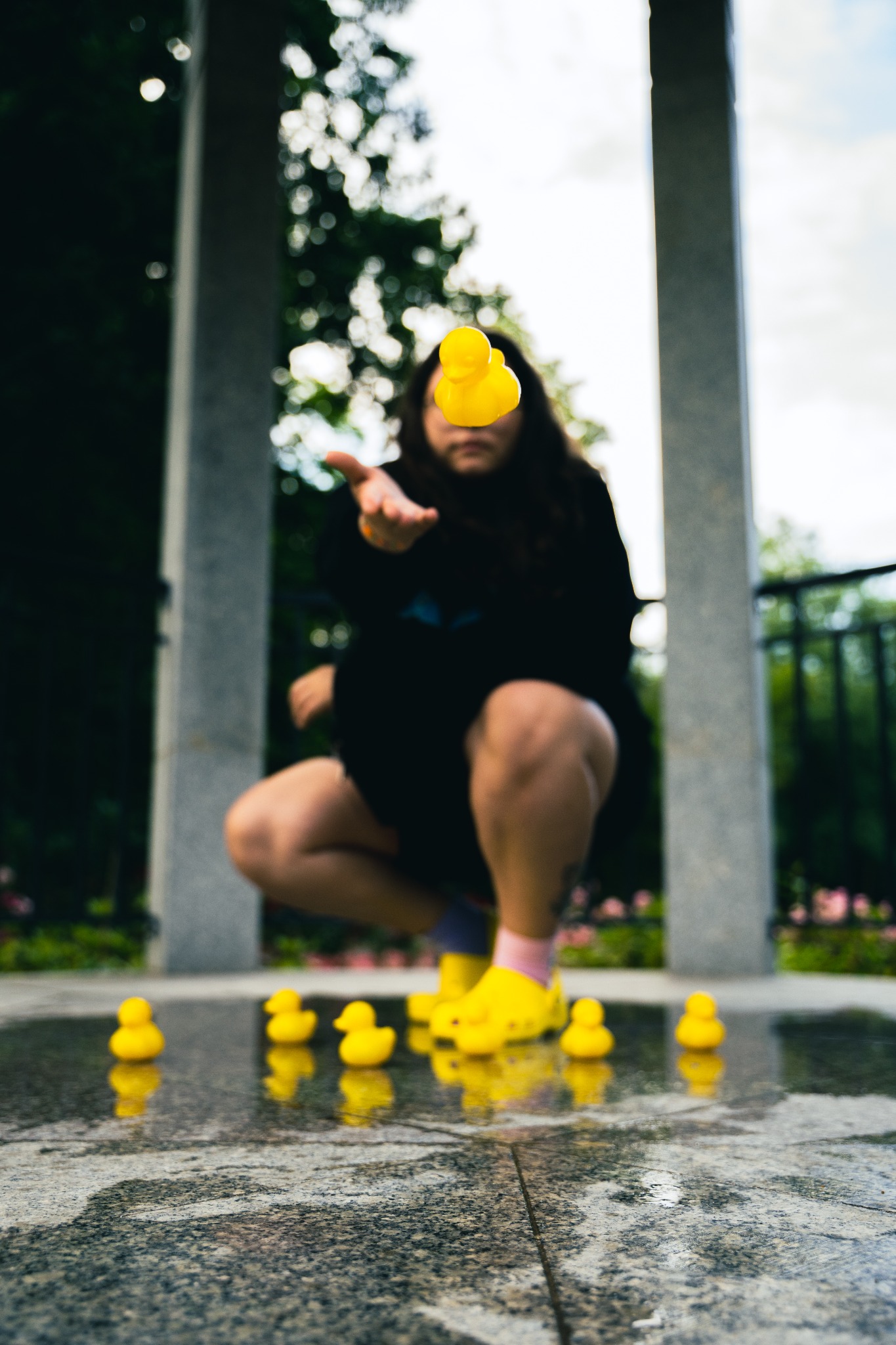 Osoba w czarnej bluzie i żółtych butach kuca na mokrej powierzchni, rzucając w górę gumową kaczkę; na ziemi leżą inne kaczki, a w tle widoczne są kolumny.