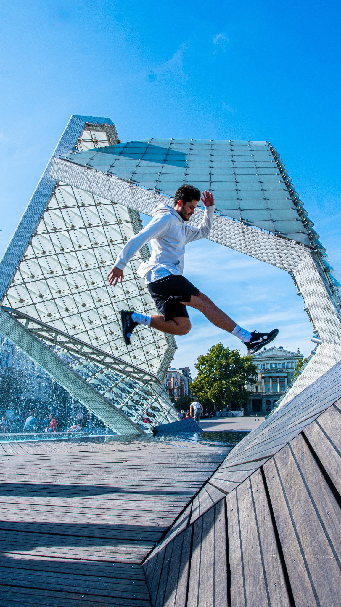 Mężczyzna w białej bluzie i czarnych spodenkach skacze przez fontannę o geometrycznej konstrukcji z elementami szkła i drewna w słoneczny dzień.