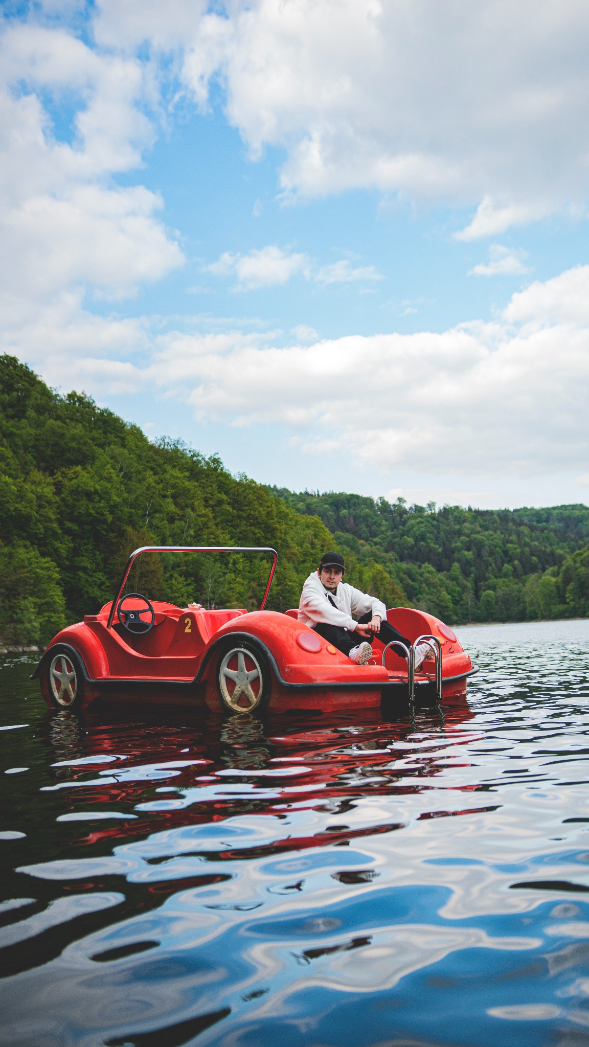 Młody mężczyzna w białej bluzie i czarnej czapce siedzi w czerwonym, samochodopodobnym rowerze wodnym na jeziorze, zalesione wzgórze w tle i błękitne niebo z białymi obłokami.