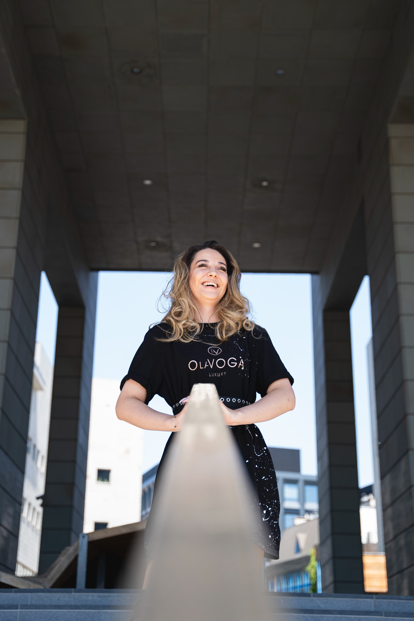 Uśmiechnięta kobieta w czarnej sukience z logo marki, stojąca na schodach z metalową barierką, w tle nowoczesna architektura miejska i błękitne niebo.