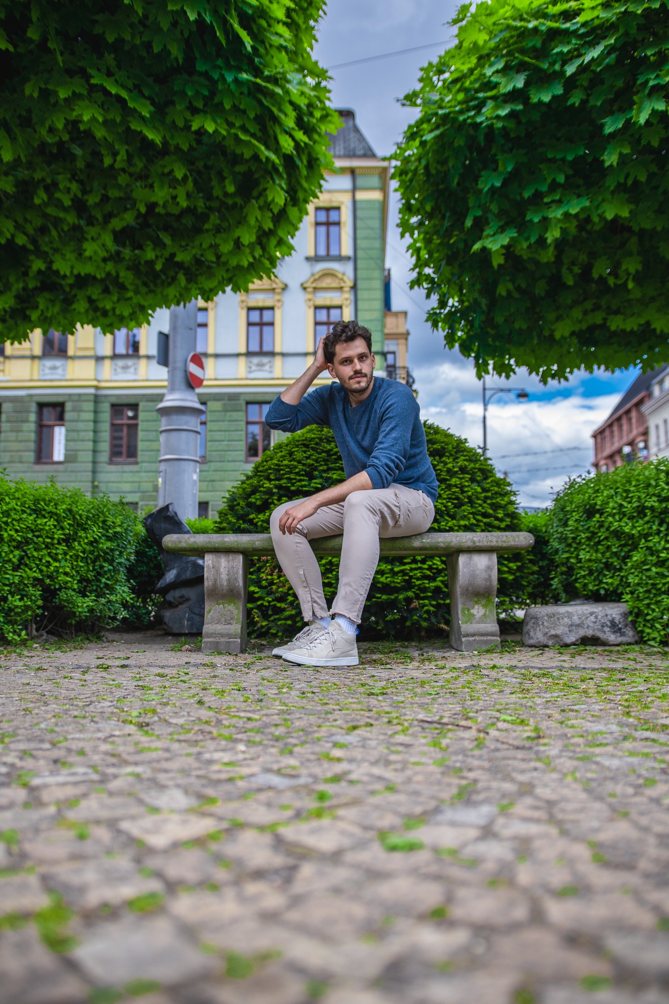 Mężczyzna z kręconymi włosami siedzi na kamiennej ławce w parku, z ręką na głowie, w tle budynek z zieloną elewacją i drzewa.