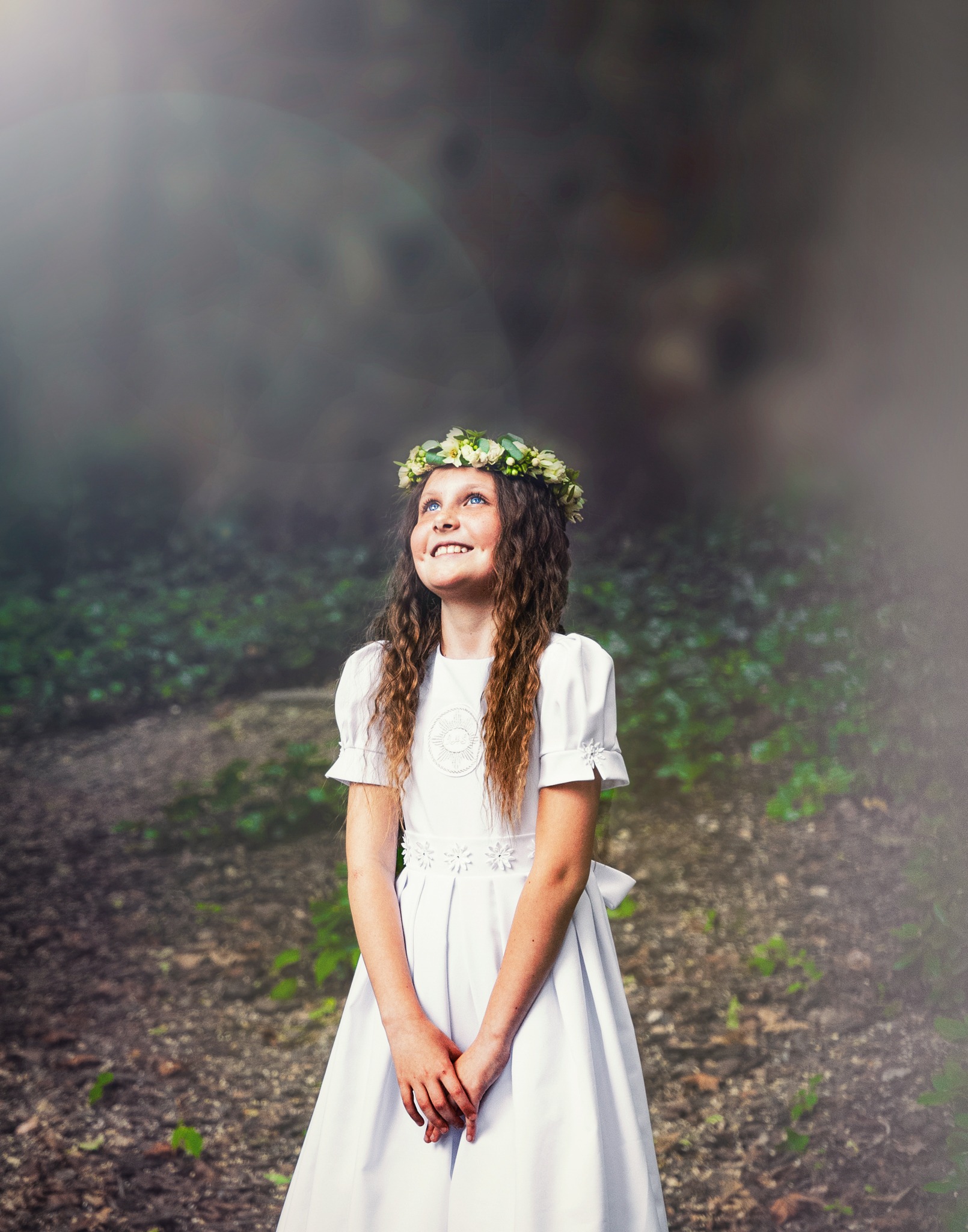 Dziewczynka w białej sukience komunijnej i wianku na głowie, spogląda w górę na tle zamazanego leśnego krajobrazu.