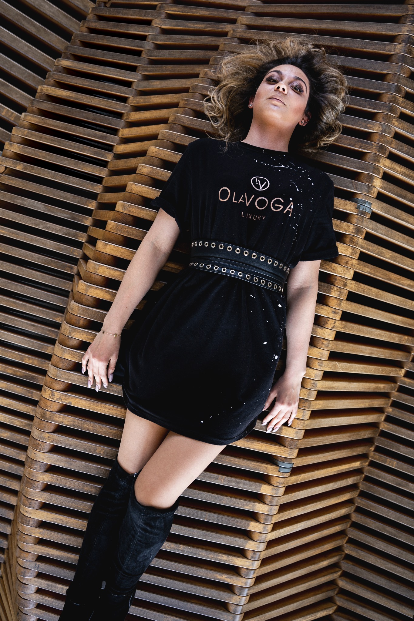 Kobieta w czarnej sukience z logo leży na drewnianej konstrukcji, pozując do zdjęcia z uniesioną głową.