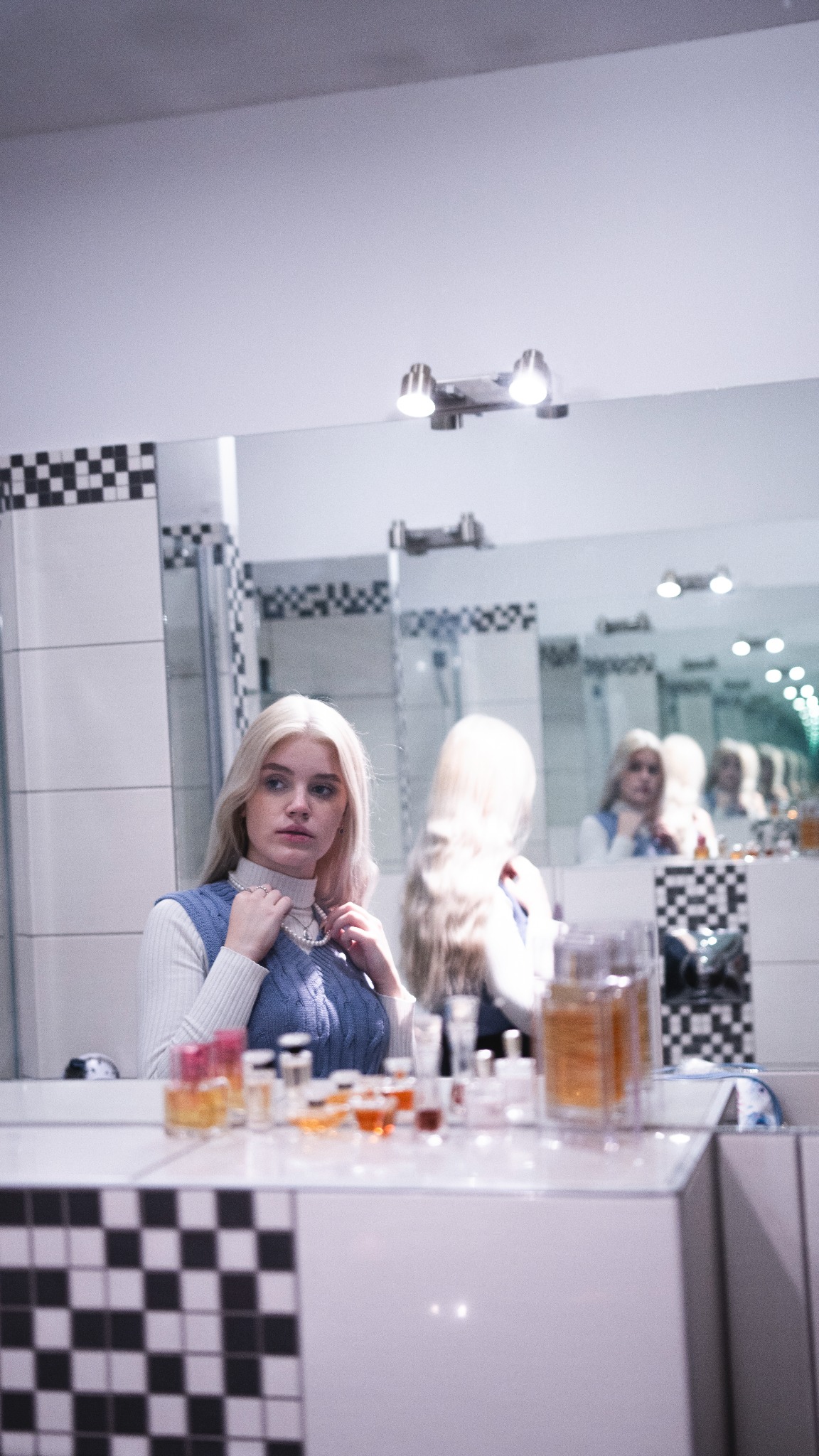 Blondwłosa kobieta w błękitnym swetrze przymierza perłowy naszyjnik przed lustrem w łazience, na blacie widoczne flakony perfum.