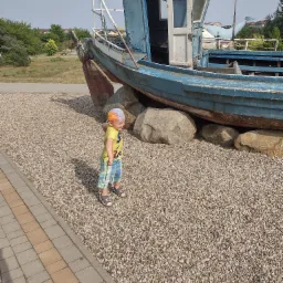 Chłopiec w żółtej koszulce i kraciastych spodenkach stoi na żwirowym podłożu obok starej, niebieskiej łodzi o numerze SPG 3978, podpartej dużymi kamieniami.