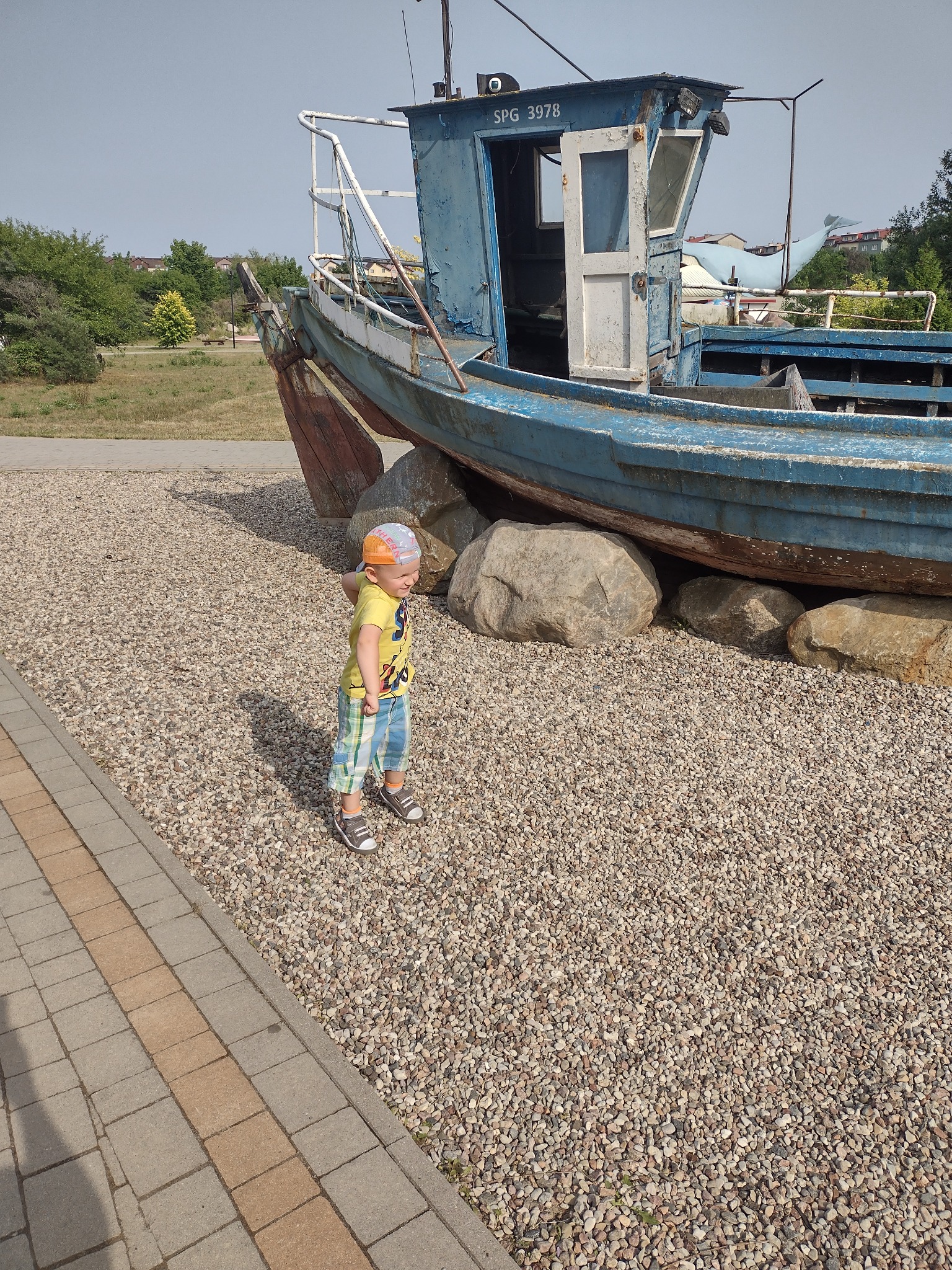 Chłopiec w żółtej koszulce i kraciastych spodenkach stoi na żwirowym podłożu obok starej, niebieskiej łodzi o numerze SPG 3978, podpartej dużymi kamieniami.