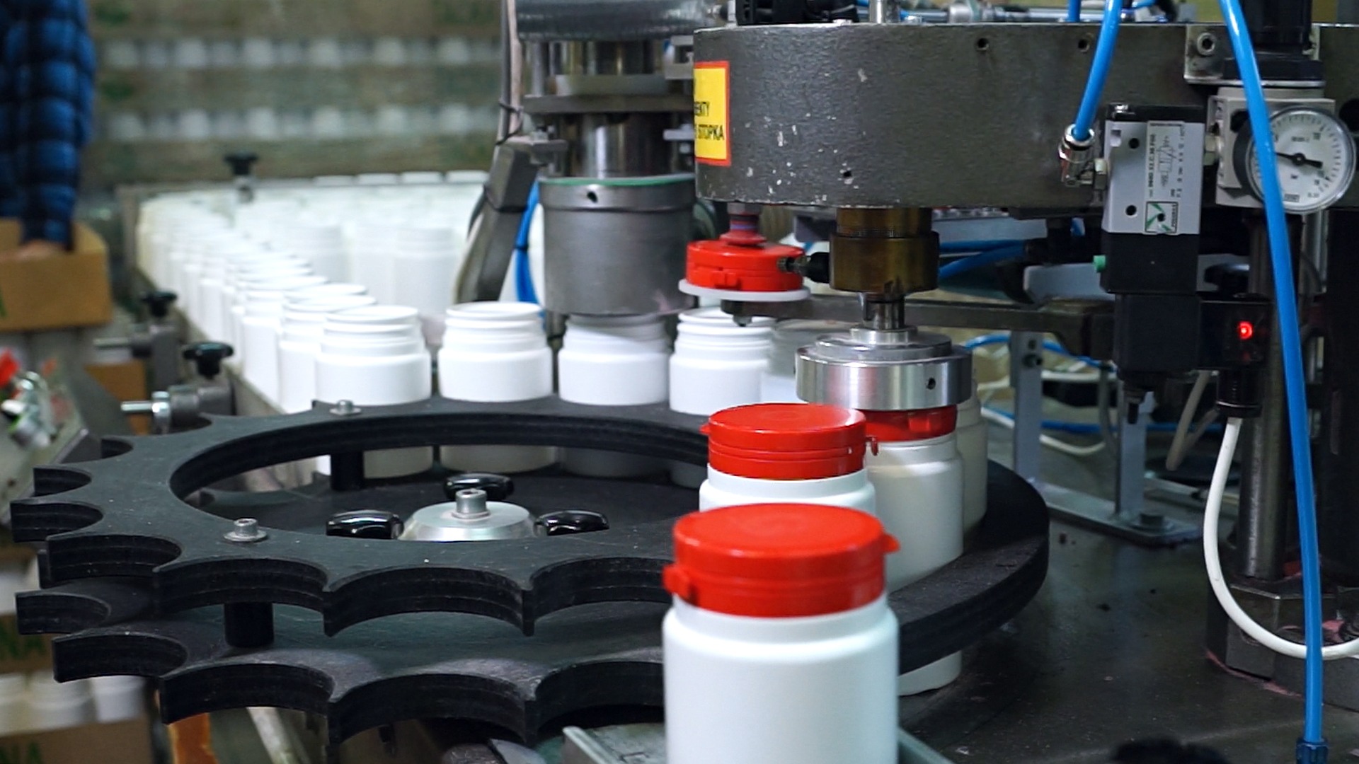Automatyczna linia produkcyjna z białymi pojemnikami z czerwonymi nakrętkami, przesuwającymi się na czarnej, zębatej tacy, podczas procesu zamykania.