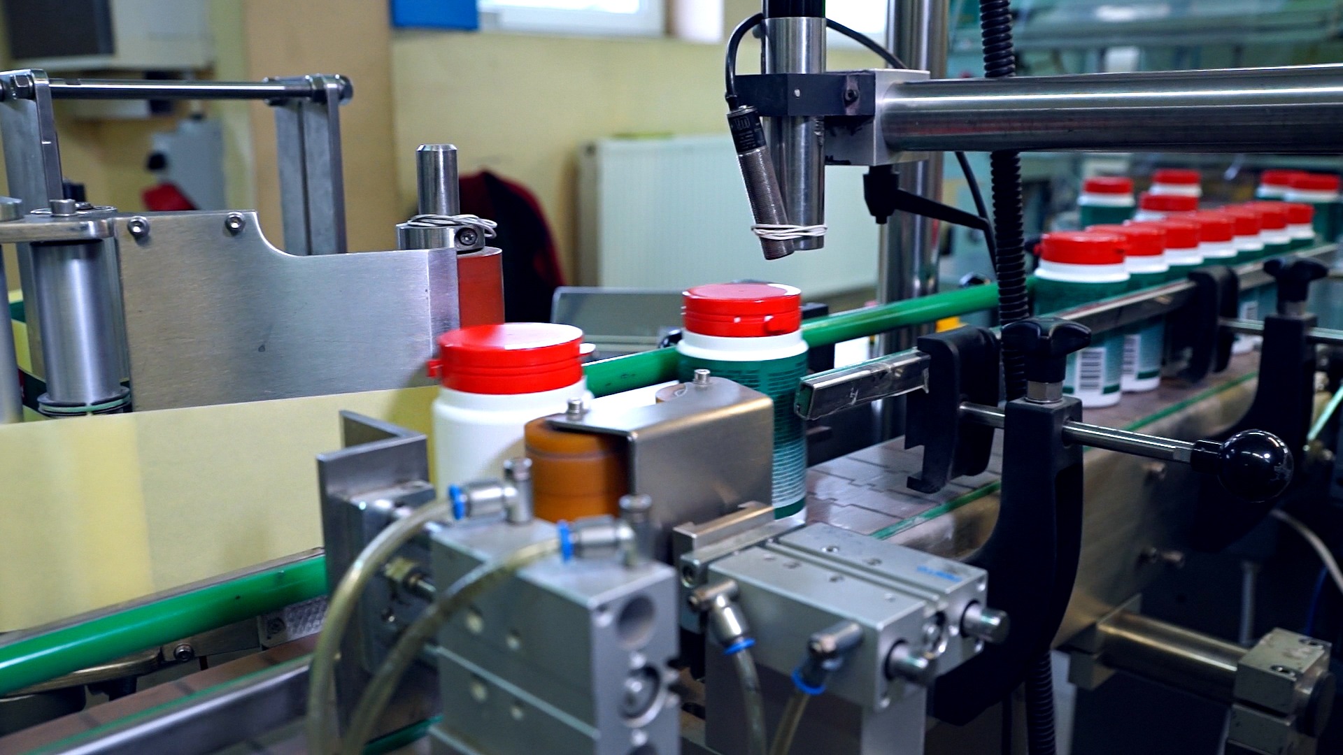 Automatyczna linia produkcyjna z pojemnikami z czerwonymi nakrętkami, prawdopodobnie preparatów biobójczych, przesuwającymi się po taśmociągu; widoczne elementy maszyn i czujniki.