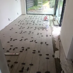 Układanie płytek podłogowych w Krakowie, z użyciem czarnych krzyżyków dystansowych, widoczny poziomica SOLA oraz wiadro z klejem PLUS3, w tle okno z widokiem na ogród.