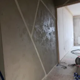Ściana z nałożoną szarą gładzią strukturalną, wyznaczającą geometryczne wzory za pomocą taśmy malarskiej, widok na korytarz w trakcie prac wykończeniowych.