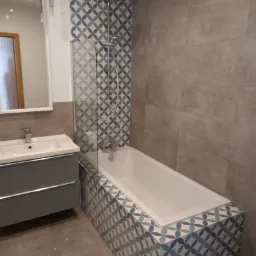 Wykończona łazienka z wanną obudowaną mozaiką w geometryczny wzór, szare płytki na ścianie, biała umywalka z szarą szafką i lustro.