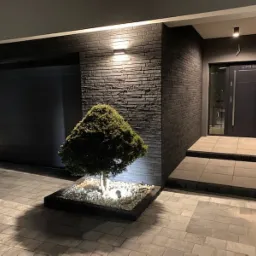 Nowoczesny dom z elewacją z ciemnej cegły, oświetlony punktowo, z równo przyciętym drzewkiem iglastym na tle kostki brukowej i wejścia.