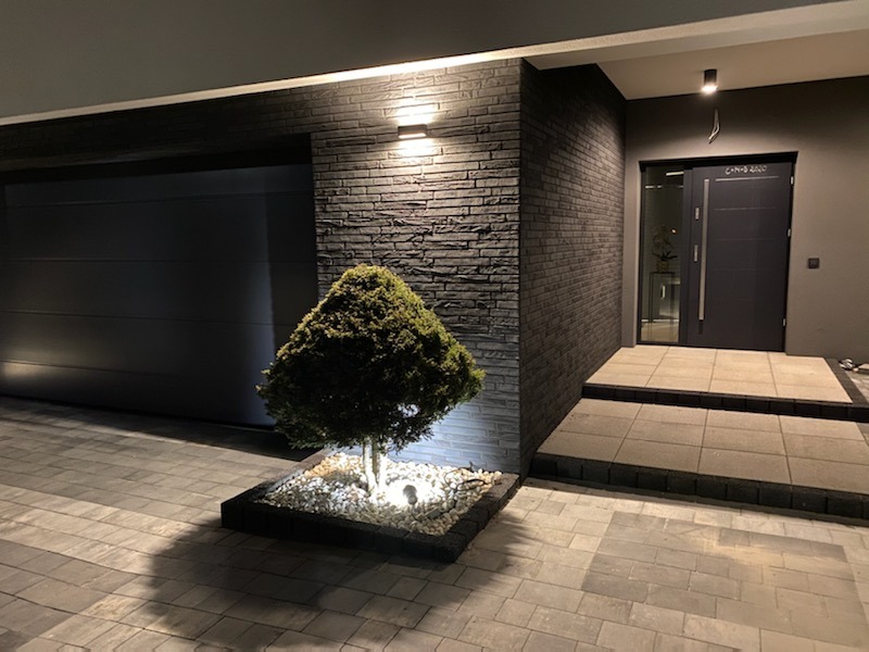 Nowoczesny dom z elewacją z ciemnej cegły, oświetlony punktowo, z równo przyciętym drzewkiem iglastym na tle kostki brukowej i wejścia.