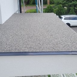 Przemysław Małasiewicz Przemix - Wykończony taras z szarej żywicy kamiennej, widoczny z góry, z obróbką blacharską w kolorze grafitowym, na tle białej elewacji domu i zielonego trawnika z samochodem osobowym w tle.