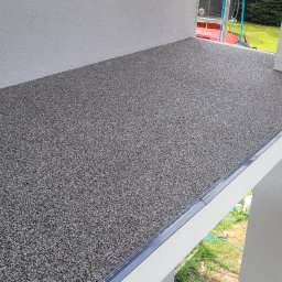 Przemysław Małasiewicz Przemix - Nowoczesny taras z szarej, drobnoziarnistej nawierzchni żywicznej, wykończony listwą przypodłogową, widok z rusztowania na tle zielonego ogrodu.