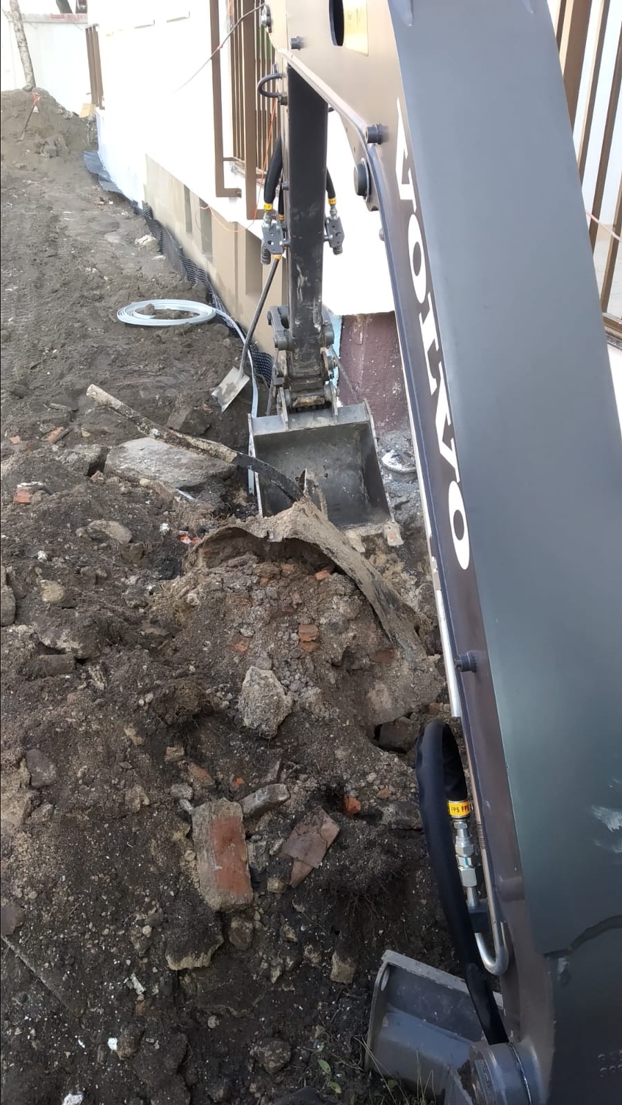 Koparka Volvo podczas prac ziemnych przy fundamentach budynku, widoczne fragmenty cegieł i rura drenarska.