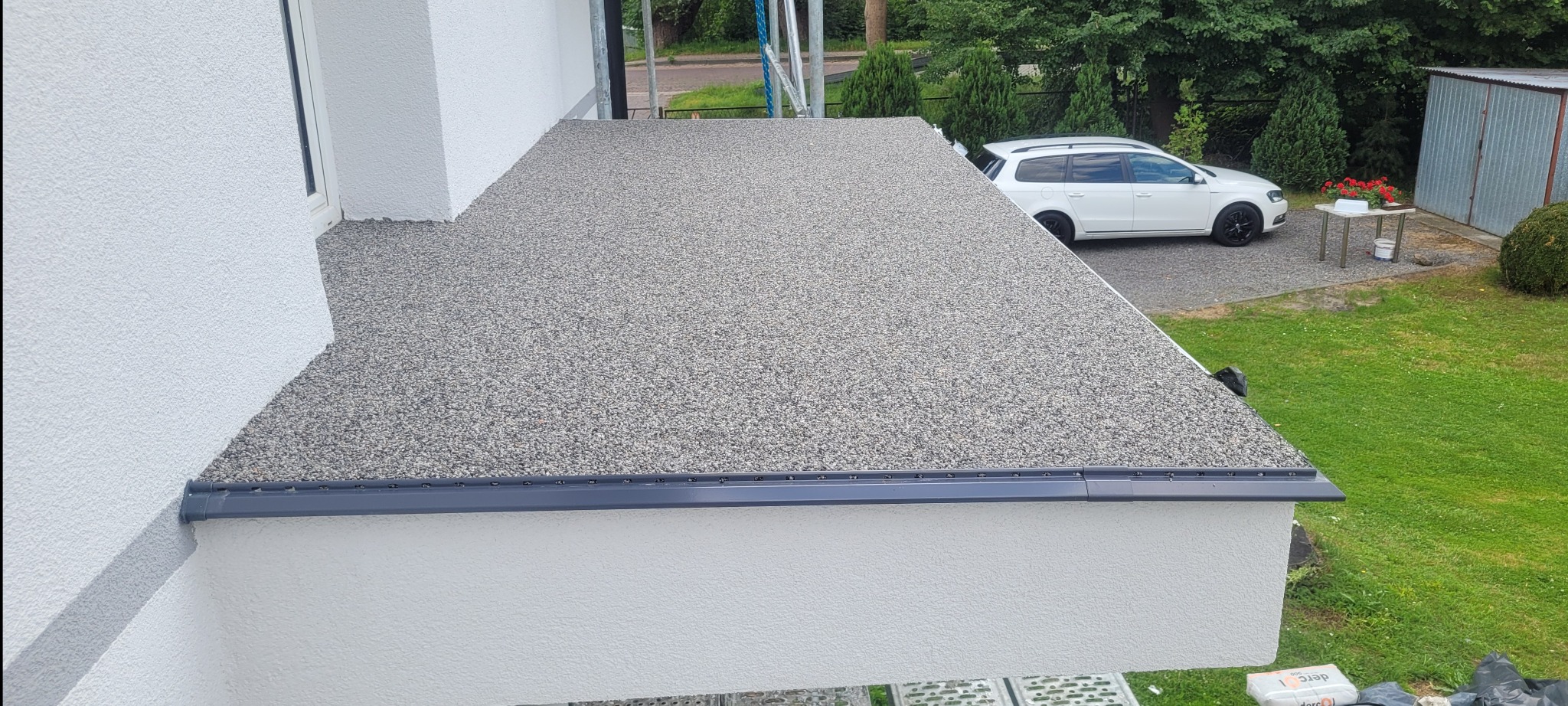Wykończony taras z szarej żywicy kamiennej, widoczny z góry, z obróbką blacharską w kolorze grafitowym, na tle białej elewacji domu i zielonego trawnika z samochodem osobowym w tle.